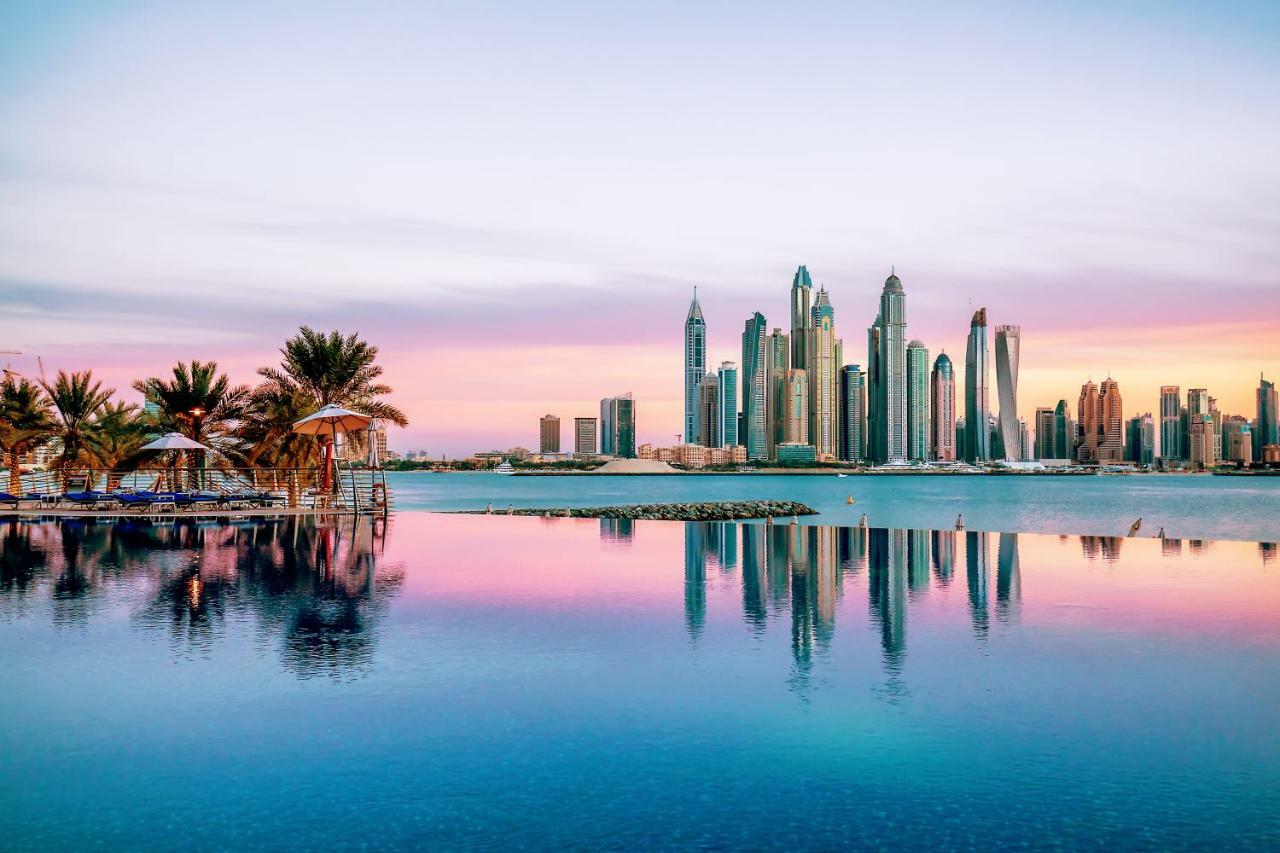 Dukes The Palm, A Royal Hideaway Hotel Дубай Екстер'єр фото The image showcases a stunning skyline view of a modern city, likely during sunrise or sunset, as indicated by the beautiful pastel colors in the sky. The foreground features a calm body of water, reflecting the skyscrapers and the colorful sky. Lush